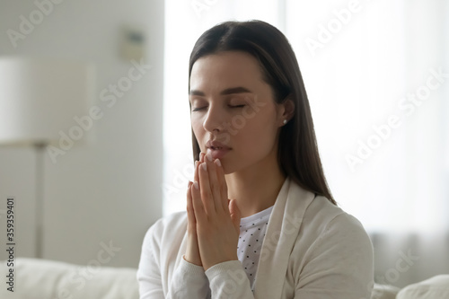 Sincere candid young woman pious deeply religious girl joined hands praying say prayer sit on sofa in living room at home. Faith in God and religion, pleading for the innermost, morning ritual concept