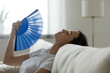 © fizkes - Close up image woman puts head on sofa cushions closed eyes feels sluggish due unbearable heat, waves hand blue fan cool herself, hot summer flat without air-conditioner climate control system concept