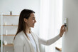 © fizkes - Smiling woman standing near wall-mounted device adjusting degrees in living room set comfortable temperature using thermostat home heating system. Owner of modern smart house, energy saving concept