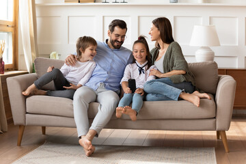 Wall Mural - Happy parents with son and daughter relaxing on couch, little girl holding smartphone, taking selfie with mother, father and brother, playing mobile device game, family enjoying weekend