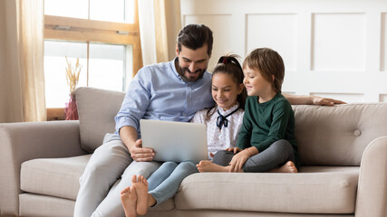 Loving father with little son and daughter using laptop, sitting on cozy couch, looking at screen, playing video game, watching video or shopping online, family spending leisure time together