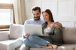 Happy young couple using laptop together, looking at screen, watching movie or video, shopping or chatting online, smiling man and woman relaxing on cozy sofa, having fun with electronic device