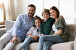 © fizkes - Family portrait happy parents with cute little son and daughter sitting on cozy couch at home, posing for photo together, looking at camera, smiling beautiful mother and father hugging kids