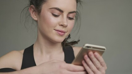 Sticker - A close-up view of an attractive smiling young woman in a black tracksuit is using her smartphone before training in a gray studio in the morning