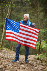 Wall Mural - full length portrait of active senior man holding american flag and smiling while enjoying hike in f