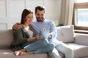 Wall Mural - Happy young couple using computer tablet together, sitting on cozy couch at home, smiling beautiful woman and man looking at screen, surfing internet, chatting online in social network