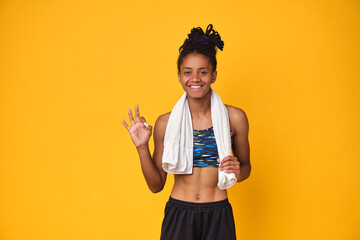 Young happy dark skinned teenager girl, athletic body isolated on a yellow background. African woman smiles, in a sports uniform holds towel on neck, shows ok sign after fitness. Copy space.