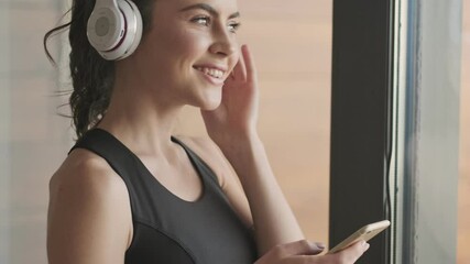 Canvas Print - A happy young woman in a black tracksuit with headphones is enjoying the music while standing near a window in a gray studio