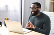 © Face Stock - Side view of African-American office employee talking via video connection while sitting at the desk. A guy using laptop for video call in contemporary office space