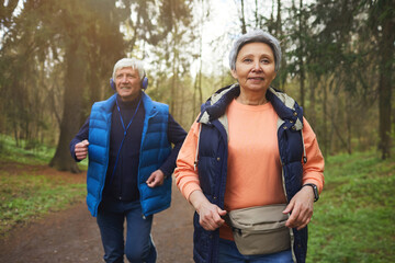 Wall Mural - waist up portrait of active senior couple running towards camera in beautiful autumn forest and smil