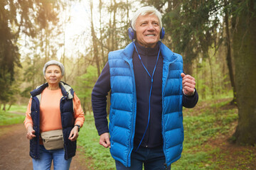 Wall Mural - front view portrait of active senior couple running in forest, focus on smiling man wearing headphon