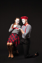 A man mime makes an offer and gives a ring  to girl mime on isolated black background 