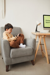 Wall Mural - Vertical full length portrait of cute boy sitting cross-legged in armchair and using smartphone, copy space