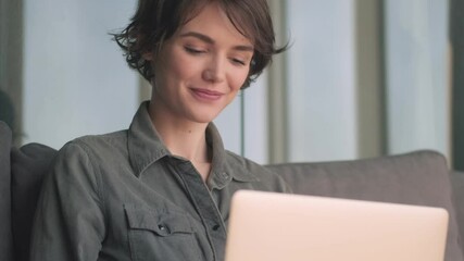 Canvas Print - Happy young pretty woman using laptop computer and looking around while sitting on sofa at home