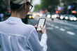 © BullRun - Cropped back view of hipster blogger holding smartphone device and touching with fingers on blank screen of device to search direction in app standing in urban setting on blurred background