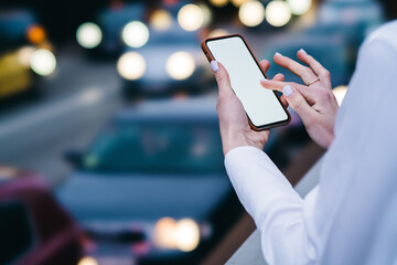 Modern smartphone with blank screen area for your internet content or website on blurred background of urban setting.Cropped view of woman's hands holding cellular and touching with finger on display