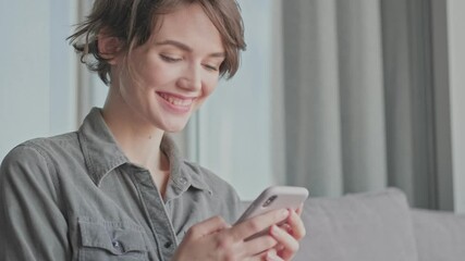 Canvas Print - Joyful young pretty woman using smartphone while sitting on sofa at home