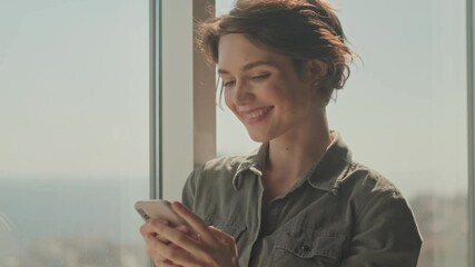 Wall Mural - Pleased young pretty woman using smartphone while standing near the window at home