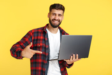 Smiling bearded hipster young man in plaid shirt pointing finger on notebook or computer over yellow background.