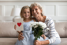 Little Granddaughter With Postcard Red Painted Heart On It Old Grandmother Holding White Flowers Sit On Sofa Pose Looks At Camera. Family Holidays Celebration, Women 8-march Day Congratulation Concept