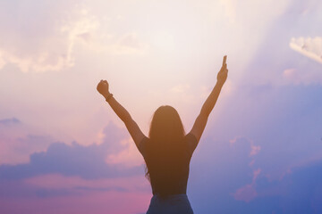 Young empowered lady raising her hands up to the sky, hand holding smart phone against sunset sky