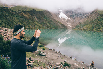 Wall Mural - Experienced male traveller in active wear travelling in mountains and making photos of exciting lake on modern smartphone.Tourist taking pictures on digital cellular during wanderlust