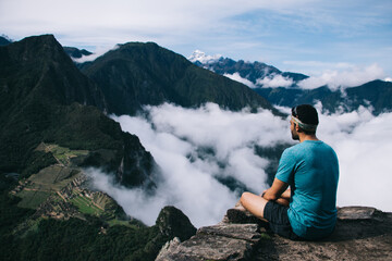 Wall Mural - Amazing scenery of high green mountains covered white clouds.Back view of young man dressed in active wear traveler resting and admiring breathtaking nature during hiking wanderlust