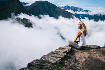 Wall Mural - Pondering blonde female tourist dressed in active wear sitting on edge of rock and thinking about calm and inspiration enjoying breathtaking view of green mountains and white clouds during trip