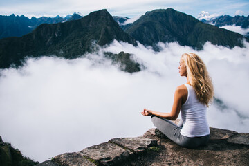 Wall Mural - Blonde young woman with blonde hair doing meditation sitting in lotus on edge of rocky hill enjoying relax time of travel.Female tourist doing yoga from high peak mountain enjoying calm of nature