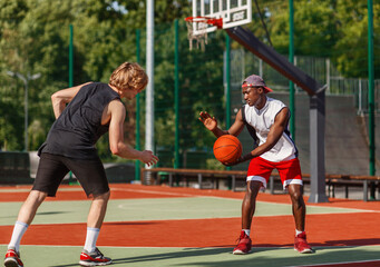 Two millennial sportsmen playing basketball at outdoor arena on summer day