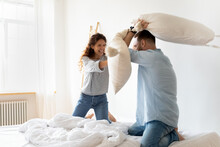 Happy Young Couple Pillows Fight And Laughing On Bed At Bedroom. Smiling Bearded Man And Woman Family Having Fun And Playing With Cushions At Home Together.