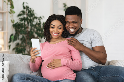 Fotografía Happy black expecting couple taking selfie at home, using phone