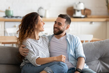 Happy Young Couple Of Man And Woman Embracing Look At Each Other Sitting On Couch In Living Room. Attractive Smiling Husband And Wife Laughing Having Fun Free Time Together.