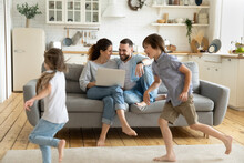 Happy Young Couple Sitting At Couch Near Running Children Using Laptop In Modern Living Room. Brother And Sister Playing In Catch Up Near Mother And Father At Home. Family Having Fun Together.