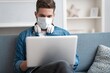 © opolja - Young caucasian man working from home, wearing protective mask, using laptop. Coronavirus pandemic, covid 19 quarantine