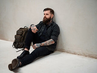 Wall Mural - Travel and education concept. Bearded hipster guy with backpack sitting on a floor in studio