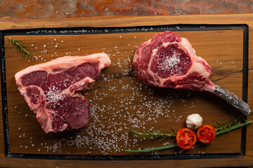 Wall Mural - Top view raw tomahawk and Tibone with bone beef steak with ingredients for grilling with a spice rub, fresh rosemary and salt , on a table with dark wood texture and space for writing