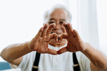 Blurred senior Asian man smiling and showing hand heart gesture for camera in cozy room at home