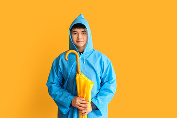 Wall Mural - Young Asian man in raincoat and with umbrella on color background