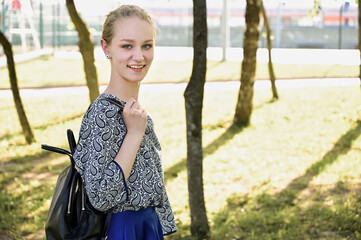 Photo of a caucasian blonde in a park. Girl stands near a tree and smiles with a backpack