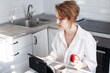 © stopabox - Happy beautiful woman with cup of coffee or tea using laptop in quarantine lockdown in the kitchen in the white shirt.