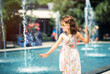 Loving life. Cheerful little girl in summer light dress and yellow sunglasses having fun playing with fountain water splashes. Summer vacation at sea. Happy child in summer hot day