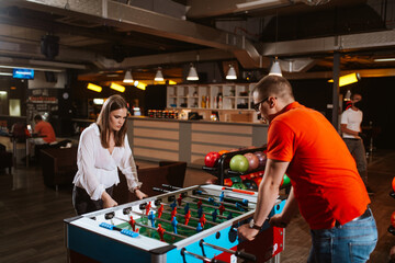 A beautiful caucasian girl and a man with glasses play table football in the playroom. Love and fun time