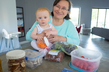 Grandmother holding red-haired baby, looking at camera. Food in containers laying on table. Portrait of senior woman sitting with grandchild. Front view. Meal prep for week concept