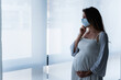 © lordn - Portrait of a pregnant woman standing by the window with medical face mask, holding her belly. Worries about childbirth during pandemic.