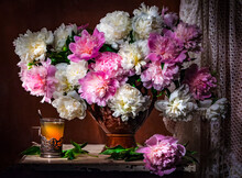 Classic Still Life With Bouquet Of Beautiful White And Pink Peonies And A Glass Of Tea In A Russian Glass Holder A Ray Of Light On Brown Background . Art Photography.