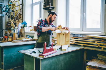 Wall Mural - serious male joiner in apron using tools for making furniture from wooden in workshop, concentrated 