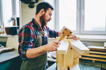 Wall Mural - pensive caucasian male carpenter in apron using hammer for making stool in his own workshop , seriou