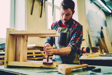 Wall Mural - skilled male carpenter in apron concentrated on work with wood and instruments owning crafts store, 