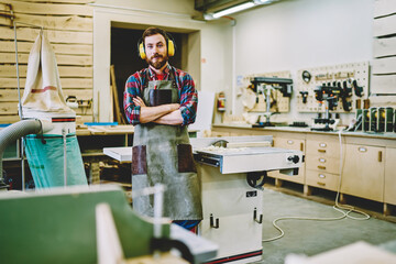 Half length portrait of self-employed woodman in work apron standing in own manufacturing fabric with crossed hands and looking at camera,bearded joiner in protective haedphones for noise cancellation